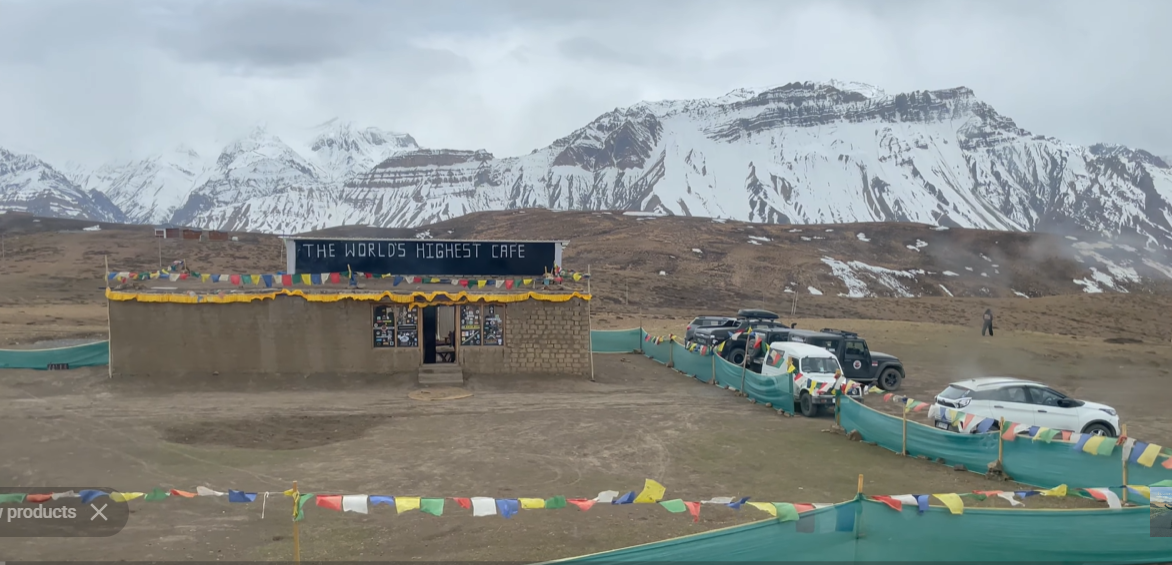 Cafe at Komic with parked vehicles, prayer flags and snowy mountains