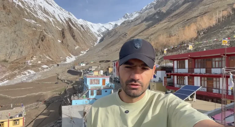 Naveen standing above Mudh village with homestays and snow-covered mountains behind him