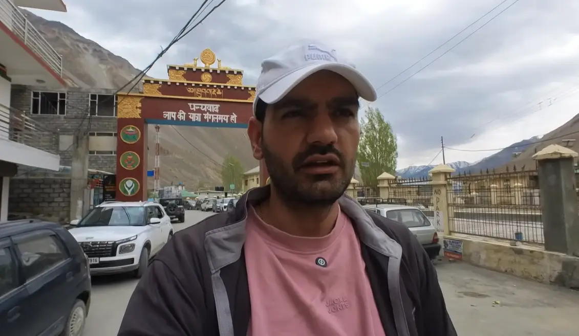 Naveen Kumar walking through Tabo village with the entrance arch and parked cars behind him.