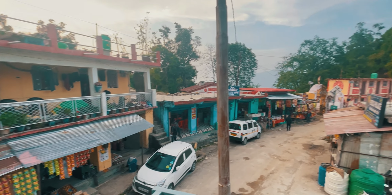Small Kanakchauri village street with shops and parked cars below the hillside
