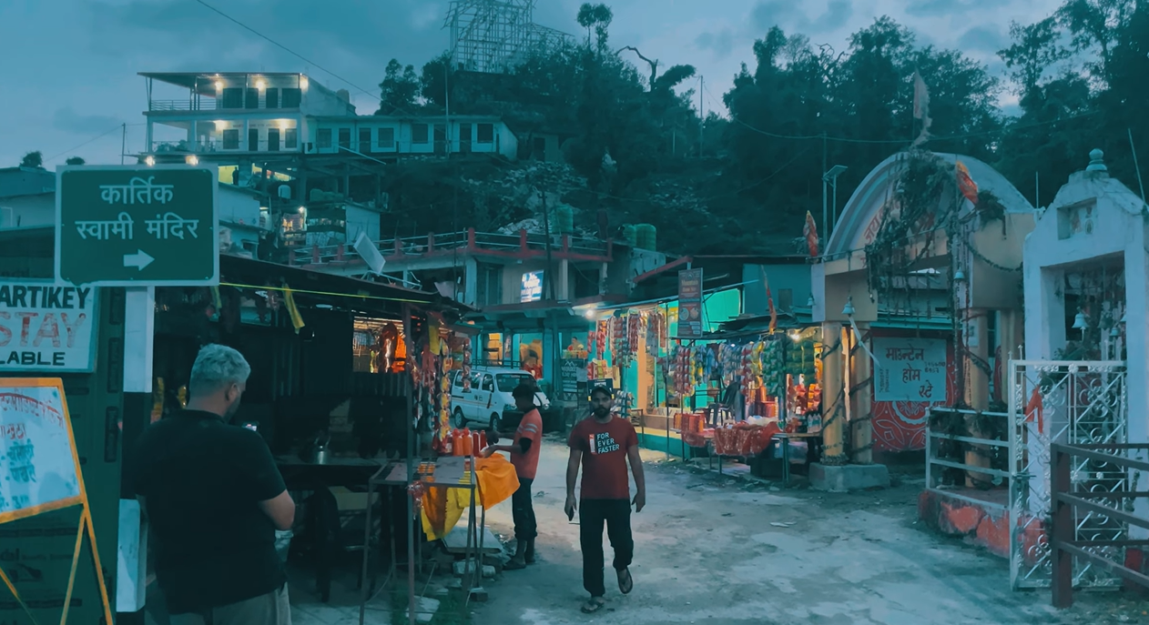 Kartik Swami Temple direction sign beside shops at the Kanakchauri trail entrance at dusk