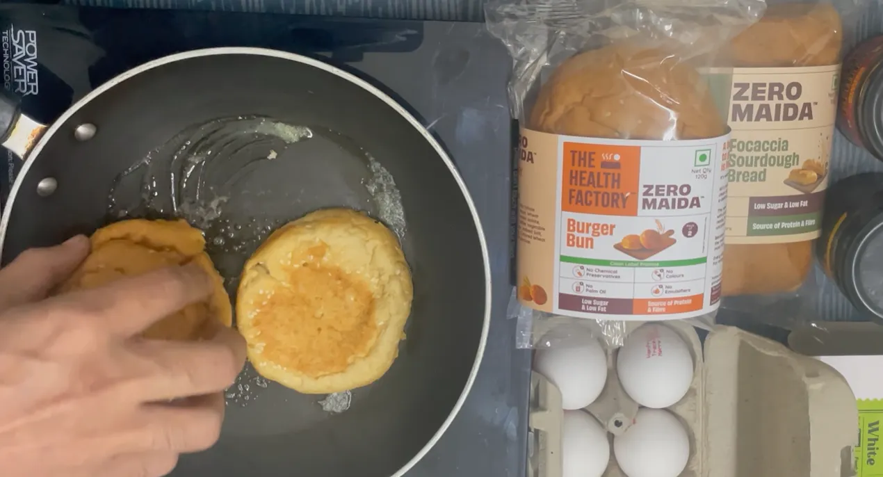 Two halves of a packaged zero maida burger bun toasting in a frying pan